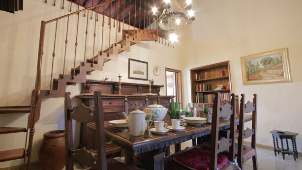 Una habitación con una escalera de madera, un candelabro, una mesa de comedor con sillas y una estantería.