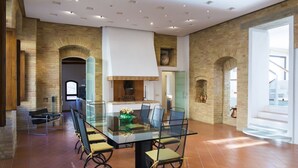 Un vaste espace dédié à la salle à manger, avec une table en verre et des chaises en métal, entouré de murs en pierre et doté d’un plafond haut avec des luminaires encastrés.
