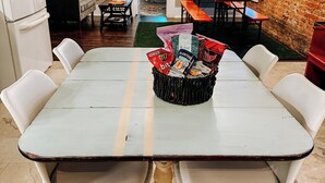 Una mesa de comedor con una cesta de aperitivos, rodeada de sillas blancas, en una habitación con pared de ladrillo y una araña.