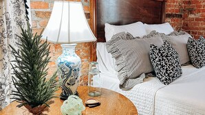 Una habitación con pared de ladrillo, cama de madera con cabecero, mesa redonda de madera con lámpara y una pequeña planta en maceta.