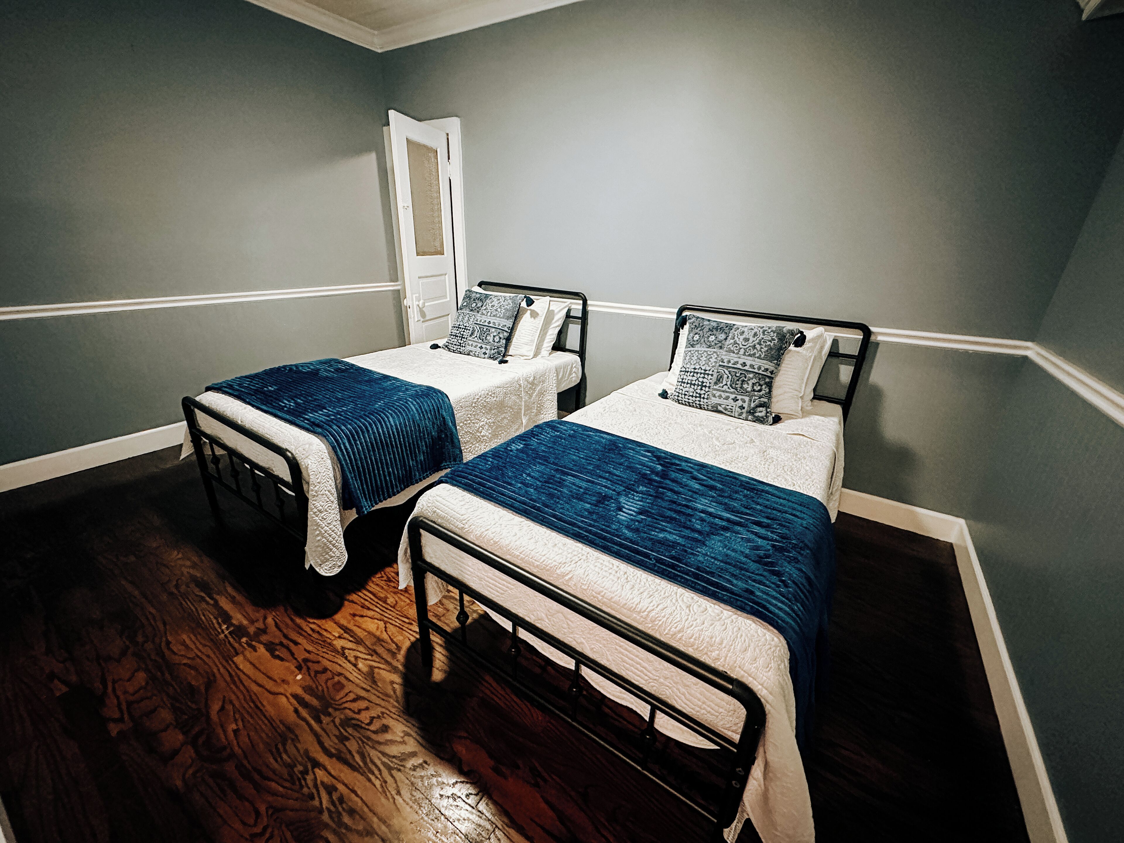 Une chambre avec deux lits simples, chacun recouvert d’une couverture bleue et agrémenté d’oreillers à motifs, un plancher en bois et une porte blanche en arrière-plan.