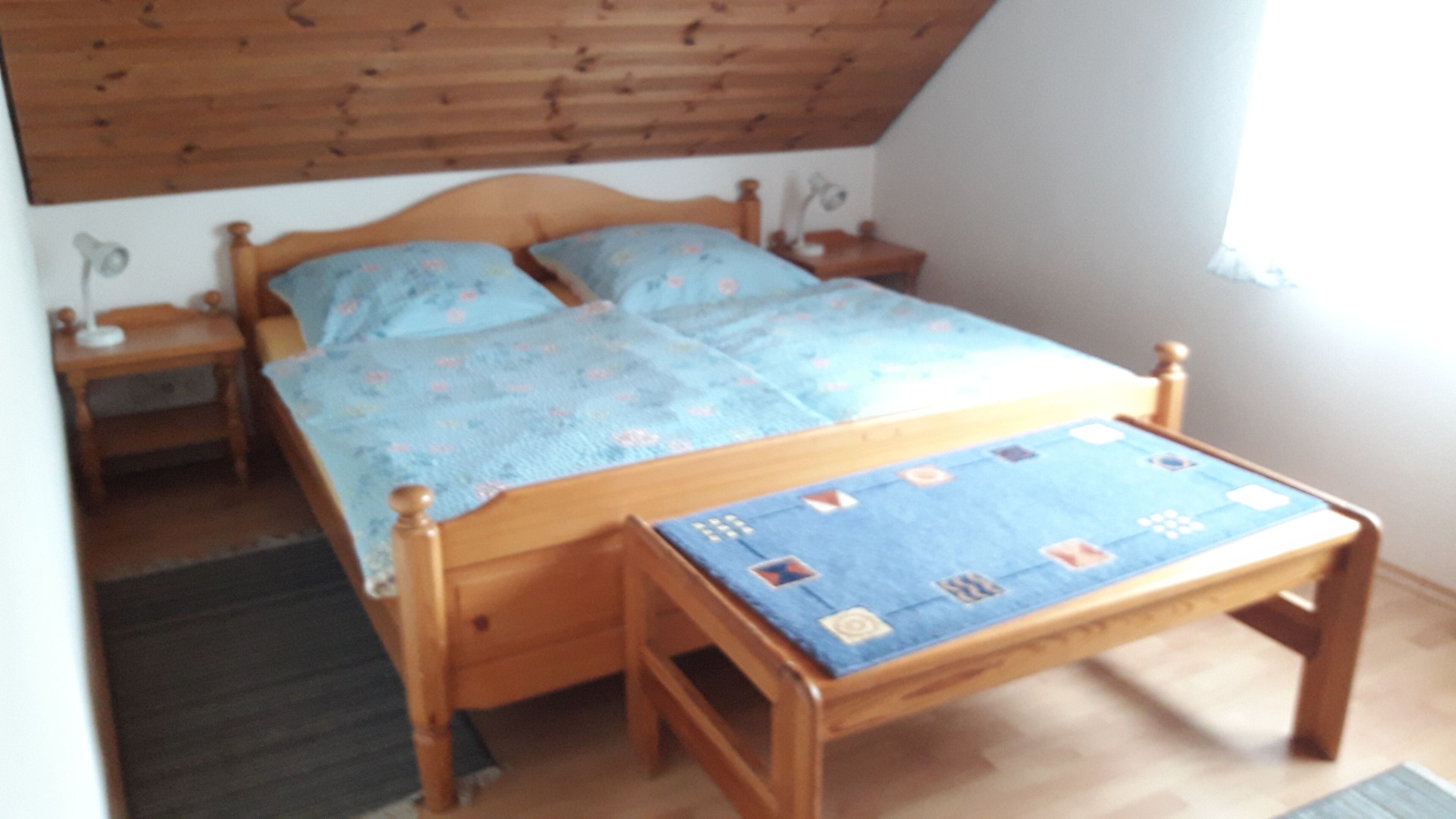 Ein hölzernes Bett mit blauer Bettdecke, ein Nachttisch, eine Lampe und ein Fenster mit Blick zum Himmel.