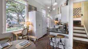 A modern kitchen with white cabinetry, a central island with stools, a dining area with a table and chairs, and a view of the outside through large windows. - Charming 2BR in Historic Savannah! (Savannah)