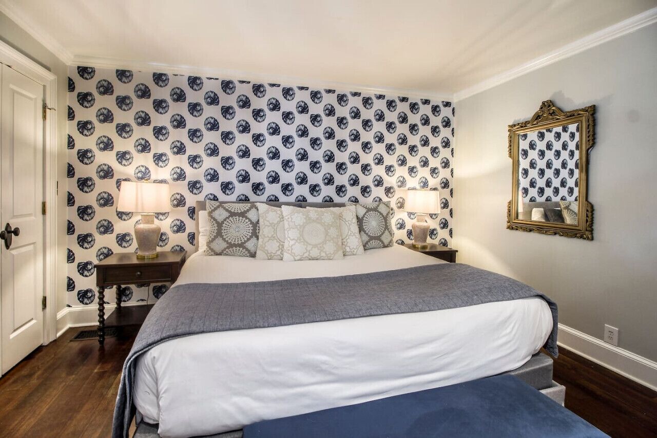 A bedroom with a large bed, two bedside tables with lamps, patterned wallpaper, and a mirror on the wall.