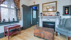 A room with a fireplace, a checkered armchair, a wooden chest, and a window with curtains. - Bristow's Inn (Township of Woolwich)