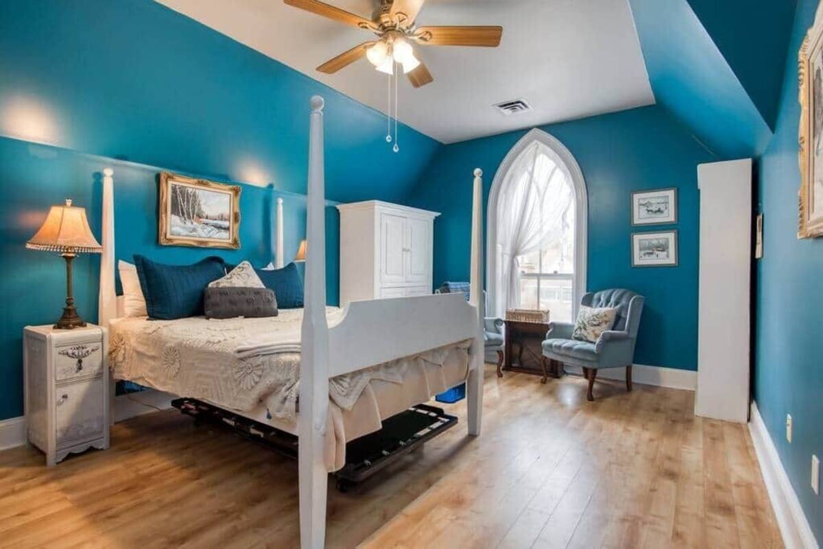 A bedroom with a four-poster bed, a ceiling fan, a window with a view, and a nightstand with a lamp.