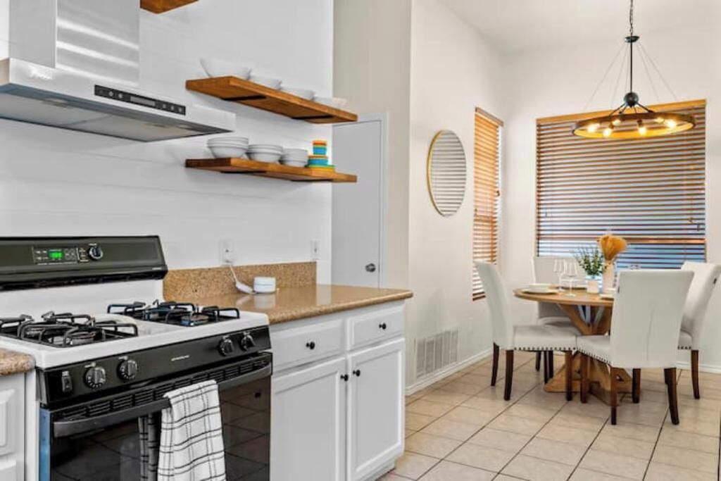 Une cuisine moderne avec des meubles blancs, une cuisinière à gaz et des étagères en bois. On y trouve également un coin repas avec une table et des chaises.