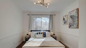 A bedroom with a large bed, a window with curtains, a chandelier, and two wall-mounted paintings. - Single-Story Charmer! 5BR! Pet Friendly Getaway

 (Indianapolis)