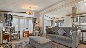 A spacious living room with a grey sofa, matching ottoman, and two armchairs. A chandelier hangs from the ceiling, and the room features large windows with patterned curtains.