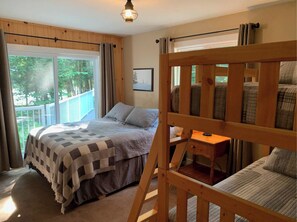 Une chambre avec un grand lit, un lit superposé en bois, une petite table et une fenêtre donnant sur un paysage verdoyant.