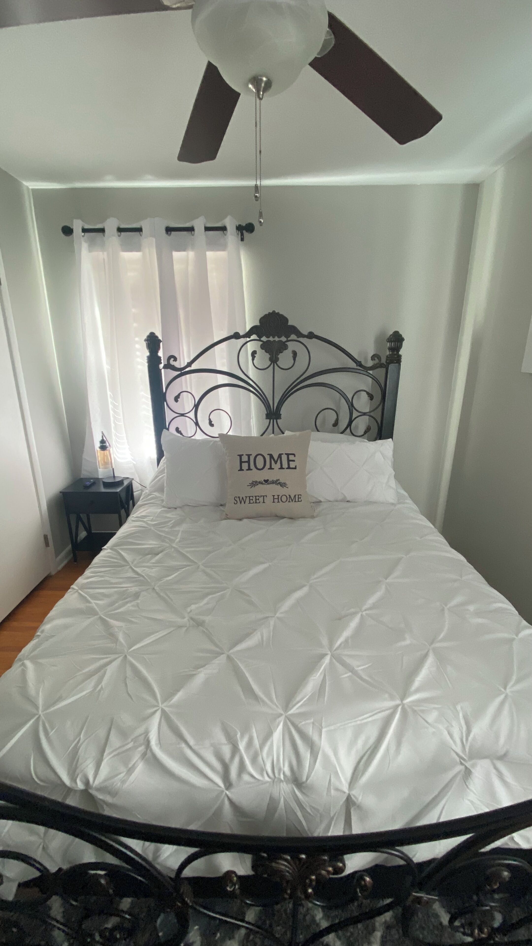 Un lit bien fait, avec un oreiller orné de l’inscription « HOME SWEET HOME », dans une pièce dotée d’un ventilateur de plafond et d’une petite table avec une lampe.