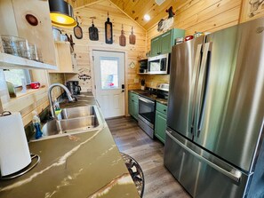 A compact kitchen with a stainless steel refrigerator, a microwave, and a stove. There is a sink with a faucet, and wooden walls with a window. - A Tyme to Chill (Sevierville)