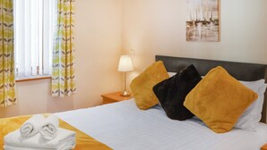 Una cama bien hecha con ropa de cama blanca y amarilla, una lámpara en la mesita de noche y un cuadro en la pared.