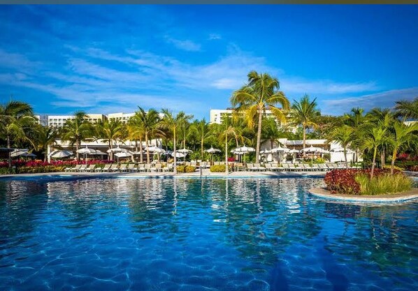 A swimming pool with lounge chairs and umbrellas, surrounded by palm trees and buildings.
