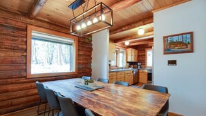 A wooden cabin interior with a large dining table, chairs, a kitchen area, and a painting on the wall.