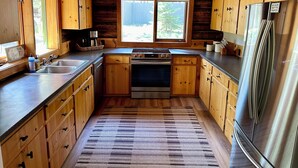 A wooden cabin-style kitchen with a large window, wooden cabinets, a stainless steel refrigerator, and a built-in oven.