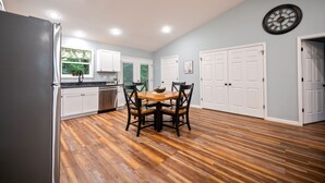A kitchen with wooden flooring, white cabinets, a dining table with chairs, and a clock on the wall. - Red River Gorge, Hollerwood Stays! High Speed Wi-Fi, Pet-Friendly! New Home! (Clay City)