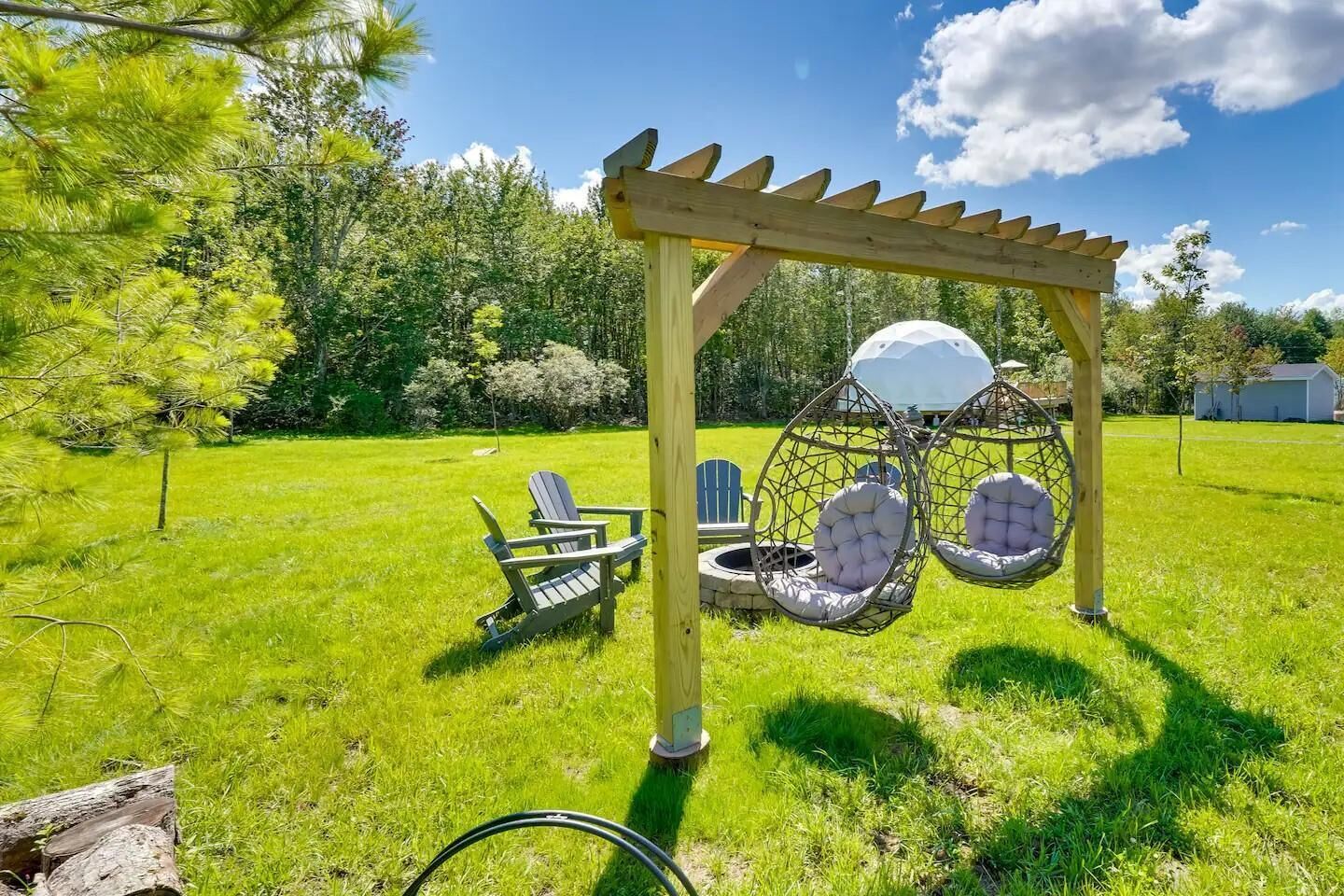 A wooden pergola with two hanging chairs and a fire pit in a grassy backyard.