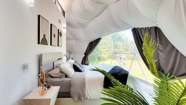 Ein modernes Schlafzimmer mit Bett, Nachttisch und Blick auf Grün durch große Fenster.