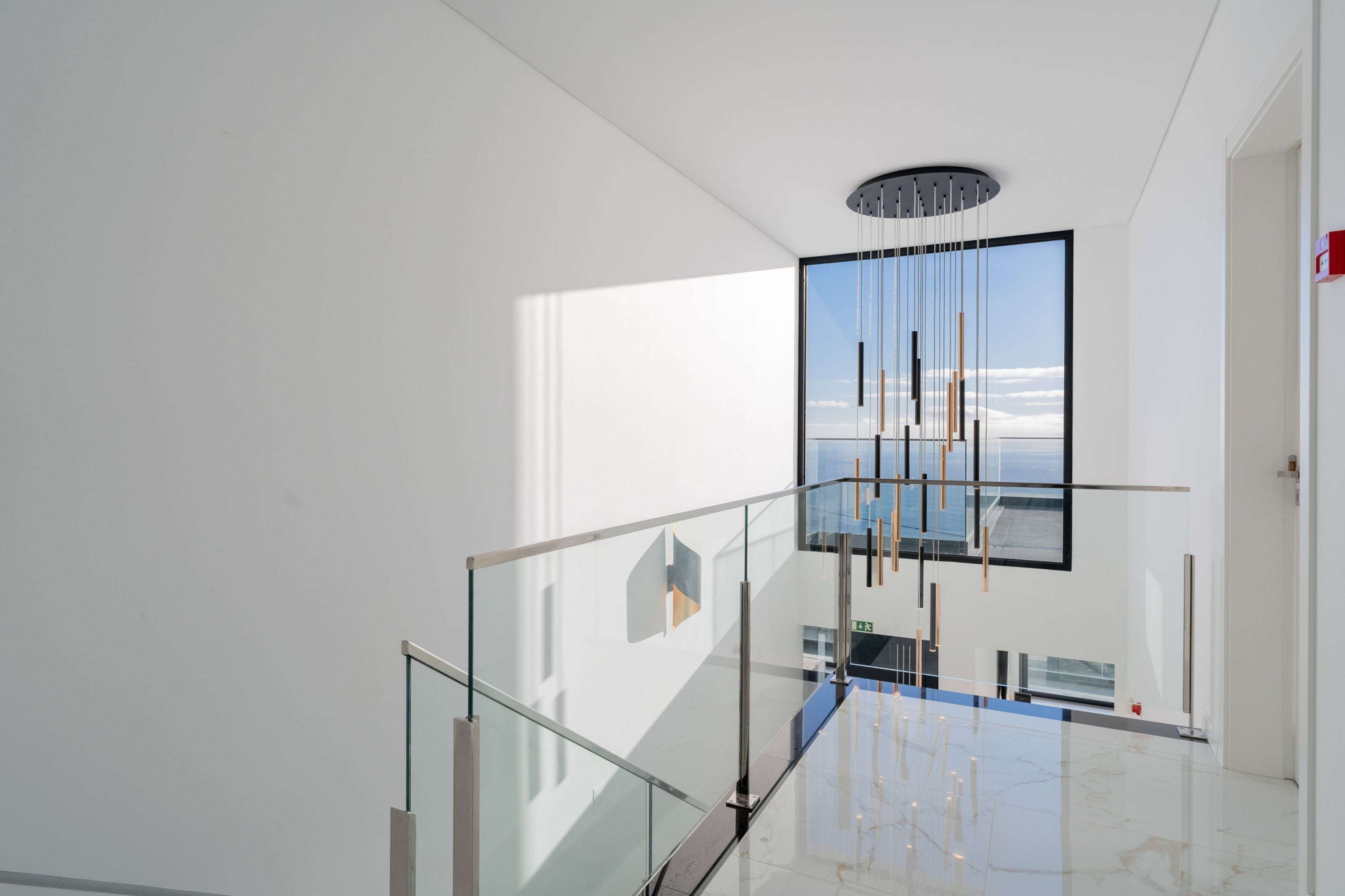 Un interior moderno con barandilla de vidrio, lámpara colgante y un amplio ventanal con vistas al cielo y nubes.