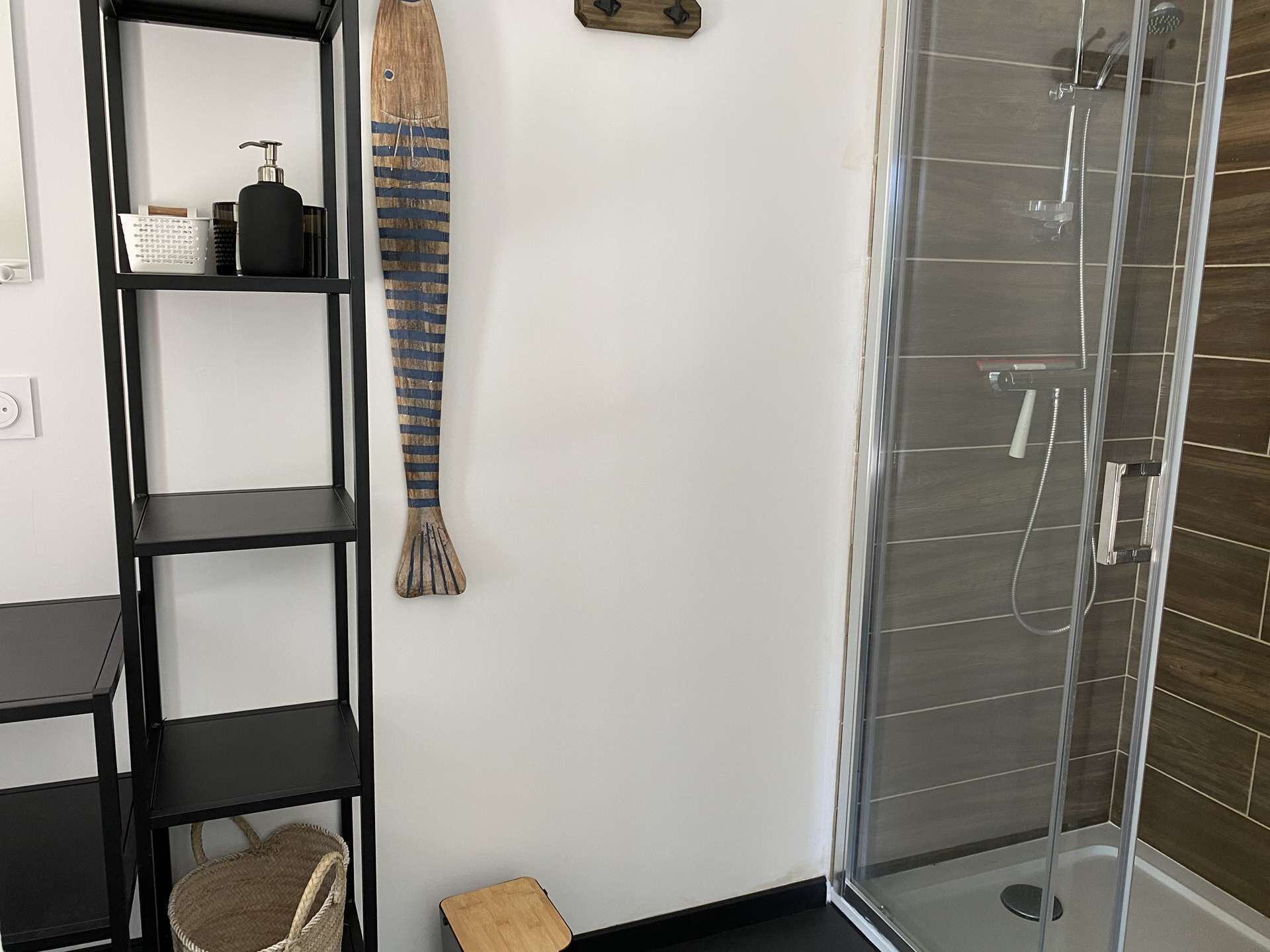 A modern bathroom with a glass shower enclosure, a black metal shelf unit with toiletries, and a woven bath mat.