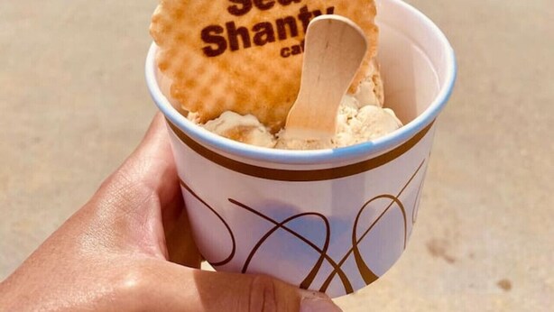 A hand holding a cup with a waffle cookie and a wooden spoon, with a beach and people in the background.