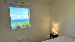 Un dormitorio con una cama, una ventana con vistas al océano, una mesita de noche con una lámpara y un calentador fijado a la pared.