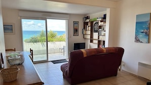 Una sala de estar con sofá, una mesa de comedor con sillas, una estantería y vista al océano a través de una puerta corrediza de vidrio.