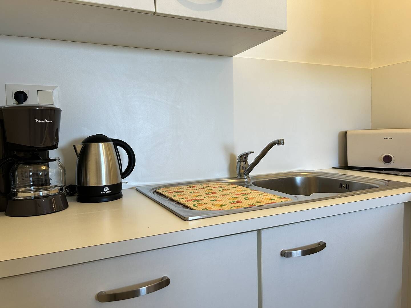 Une petite cuisine équipée d’une machine à café, d’une bouilloire et d’un grille-pain. On y trouve un évier avec un robinet et un petit appareil électroménager posé sur le plan de travail.