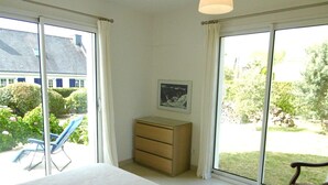 A bedroom with a bed, a wooden chest of drawers, a chair, and a large window offering a view of a garden and a building outside.