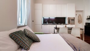 A modern bedroom with a bed, pillows, a bedside table, a window with curtains, and a kitchenette in the background. - All in One - MyHomeinComo (Como)