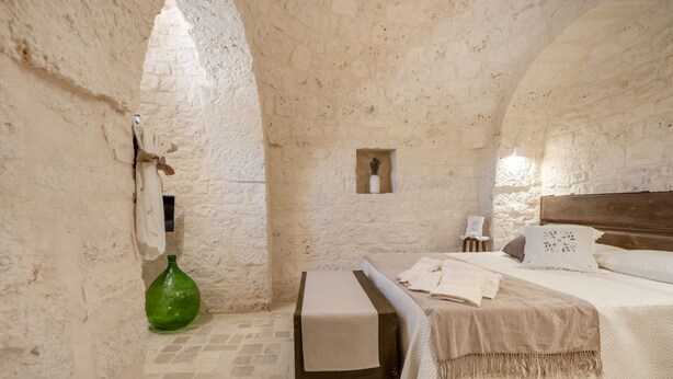 A rustic stone-walled room with a bed, a small table, and a green bottle. - GuestHost - Beautiful trullo with typical and evocative architecture, equipped with every comfort to accommodate up to 6 people with great comfort in 100 m2, with 2 bedrooms and 2 bathrooms. The accommodation is located in the countryside of the Itria Va (Martina Franca)