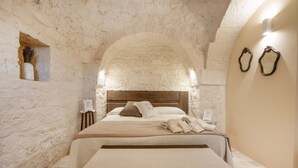 A bedroom with a stone wall, a wooden headboard, a bed with a white bedspread, and a small table with a lamp. - GuestHost - Beautiful trullo with typical and evocative architecture, equipped with every comfort to accommodate up to 6 people with great comfort in 100 m2, with 2 bedrooms and 2 bathrooms. The accommodation is located in the countryside of the Itria Va (Martina Franca)