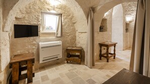 A rustic room with a stone wall, a mounted TV, a window with curtains, a small wooden stool, and a table. - GuestHost - Beautiful trullo with typical and evocative architecture, equipped with every comfort to accommodate up to 6 people with great comfort in 100 m2, with 2 bedrooms and 2 bathrooms. The accommodation is located in the countryside of the Itria Va (Martina Franca)