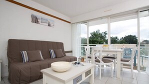 A modern living room with a brown sofa, a white table with a bowl, and a balcony with a table and chairs. - Logement à 700m de la plage, sur le golf Moliets ! (Moliets)