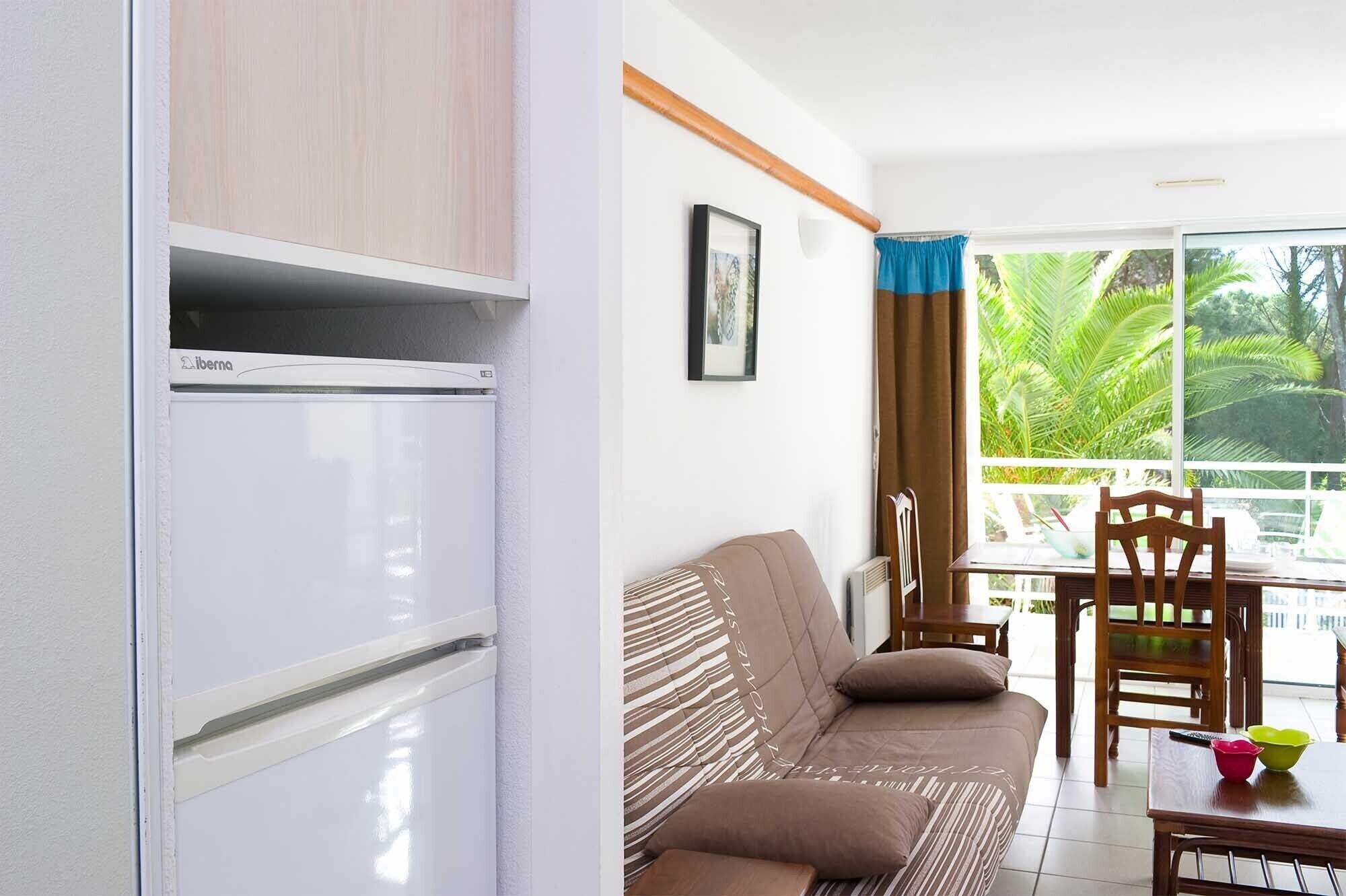 A compact living space with a kitchenette, a sofa, a dining table, and a view of greenery through the window.