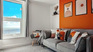 A living room with a grey sofa, a small table, and a window with a view of the sea. - Ocean View Apartment (Bridlington)