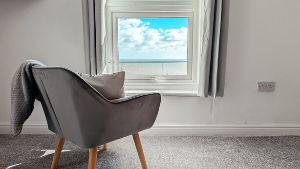 A modern room with a grey chair, a view of the sea through a window, and a carpeted floor. - Ocean View Apartment (Bridlington)