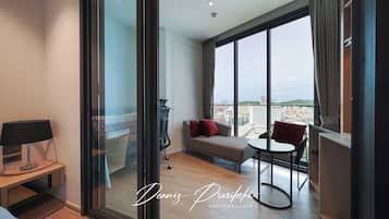 A modern hotel room with a large window, a sofa, a small table, and a desk with a lamp.
