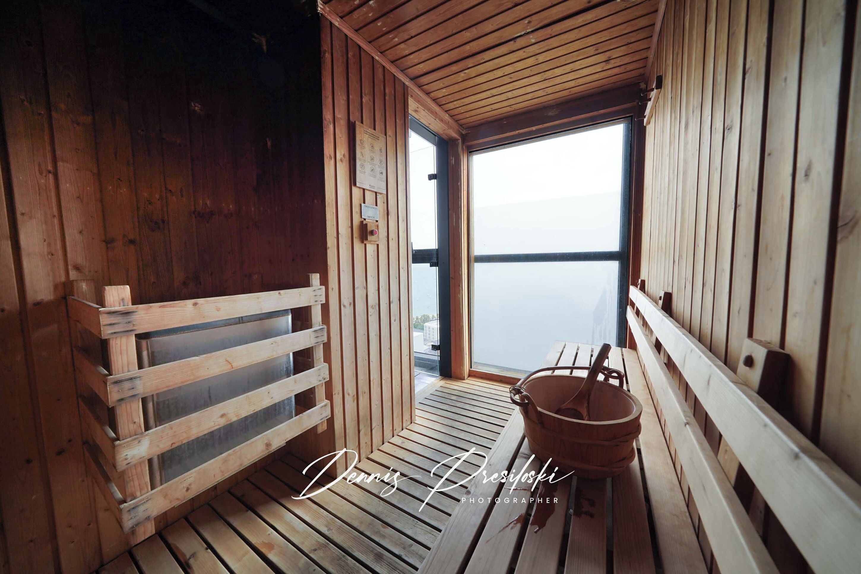 A wooden sauna room with a bench, a bucket, and a window offering a view of the water.