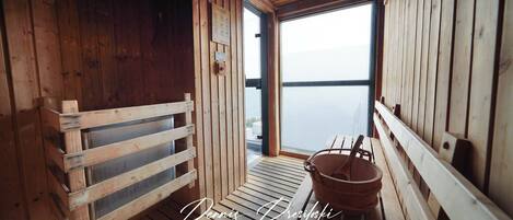A wooden sauna room with a bench, a bucket, and a window offering a view of the water.