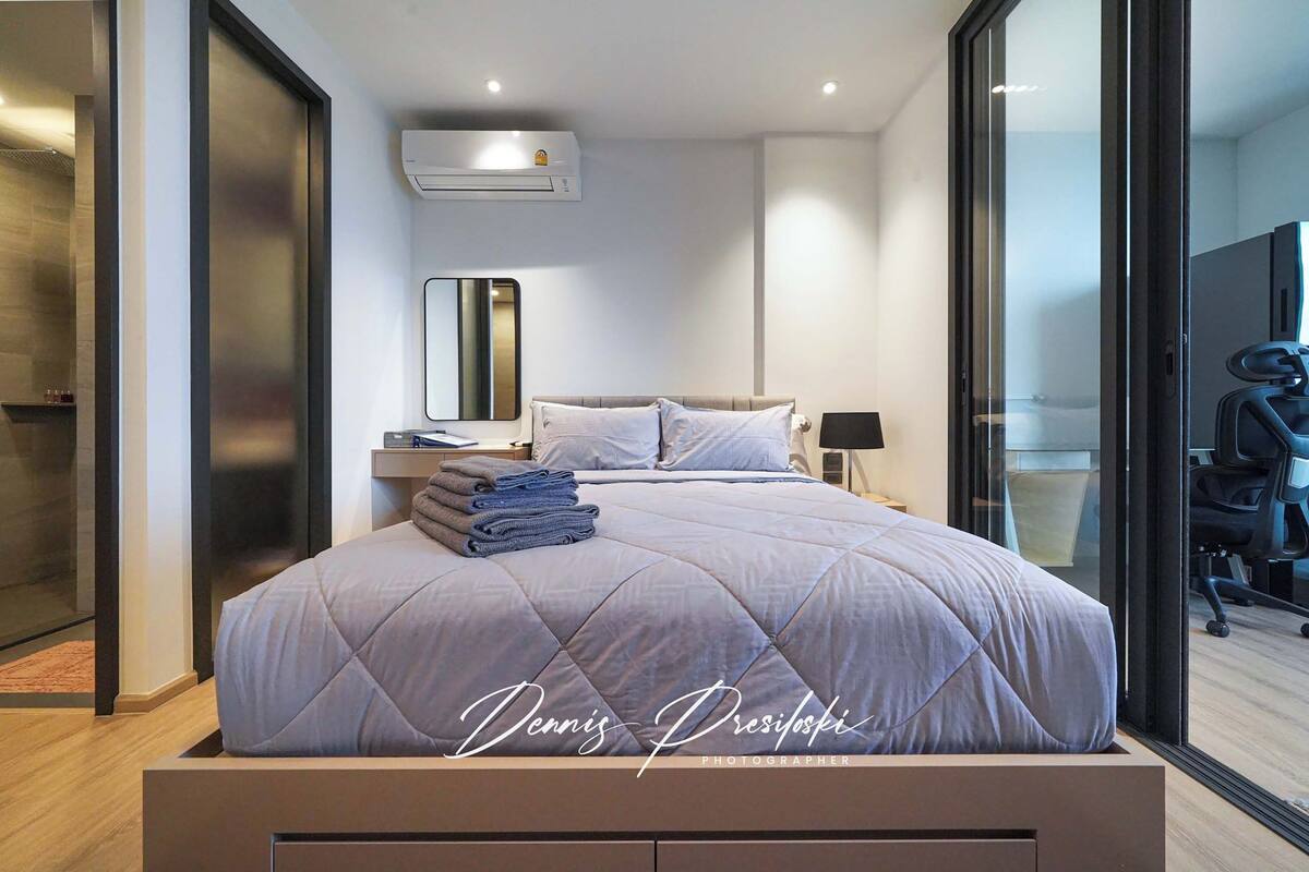 A modern bedroom with a large bed, a bedside table, a mirror, and a view through a glass door to a bathroom.