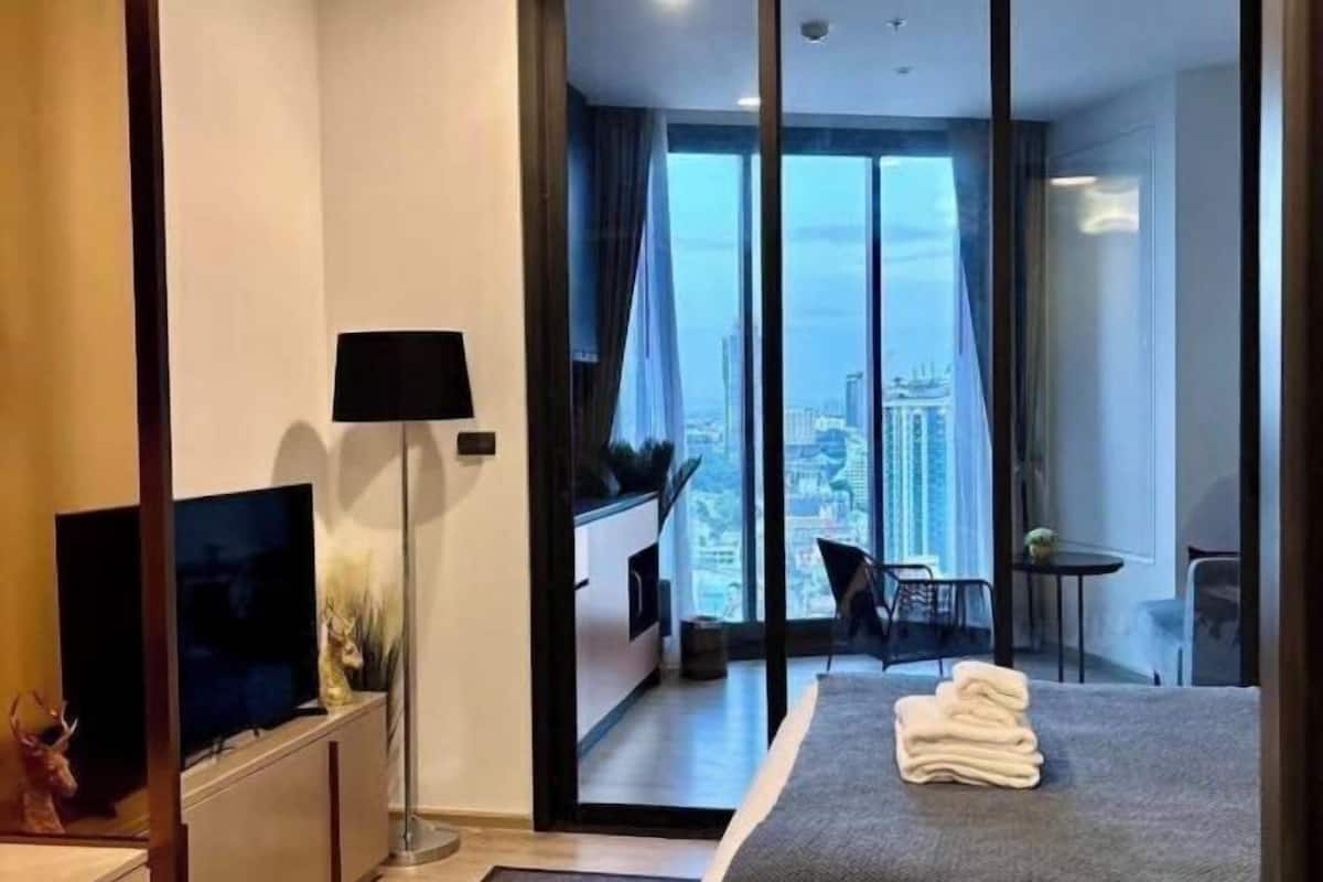 A modern hotel room with a large window offering a city view, a flat-screen TV, a lamp, and a neatly made bed.