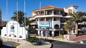 Een meerlaags hotel met balkons, een bord met appartementen te koop en palmbomen.