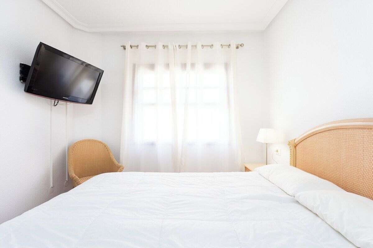 Una camera d'albergo con un letto, una televisione a parete, una sedia, un comodino con una lampada e una finestra con tende.