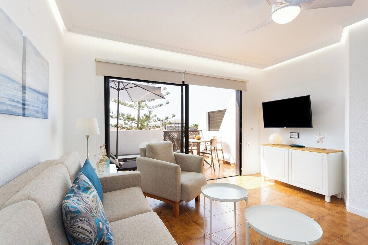 Un salon moderne avec un canapé, des fauteuils, un téléviseur et un coin repas visible à travers une porte vitrée.