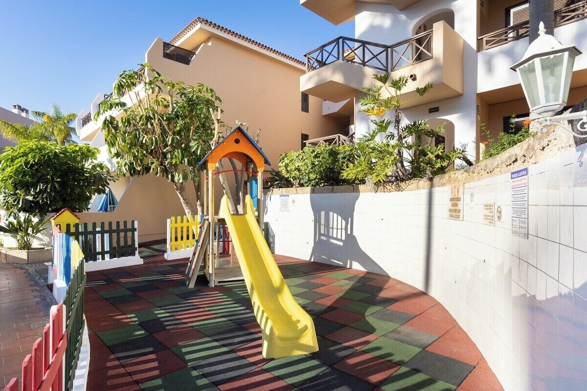 Une aire de jeux avec un toboggan jaune et une structure d’escalade, entourée d’un tapis de jeu coloré et d’un mur blanc doté d’un balcon.