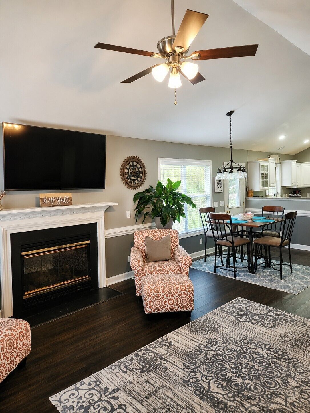A living room with a fireplace, a ceiling fan, a dining area with chairs, and a kitchen in the background.