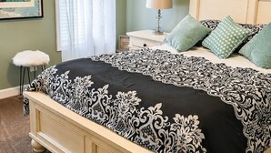 A bedroom with a bed, a nightstand, a lamp, a chair, a framed picture, and a window with curtains.