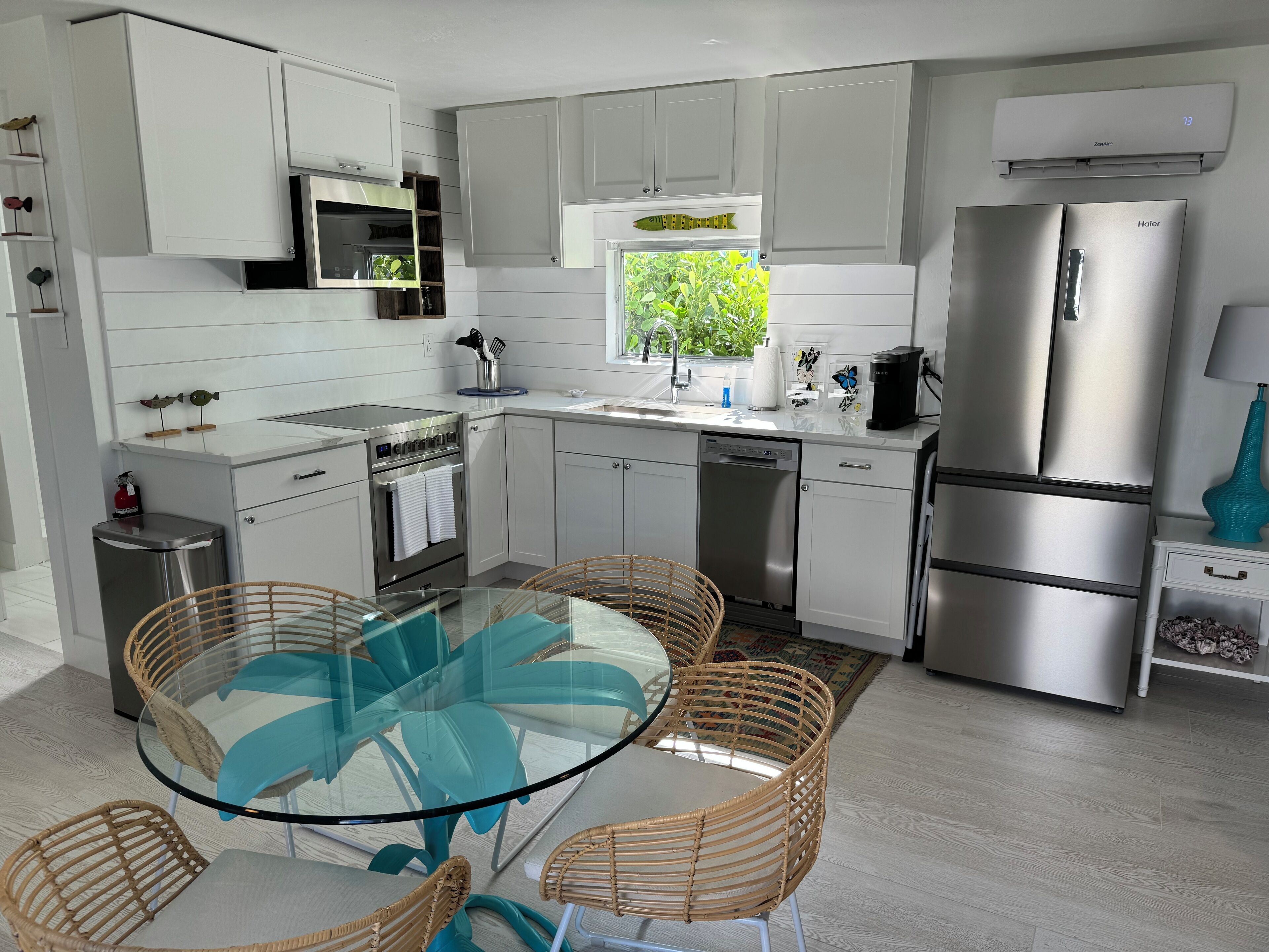 A modern kitchen with white cabinetry, stainless steel appliances, a glass dining table with wicker chairs, and a window with a view of greenery.